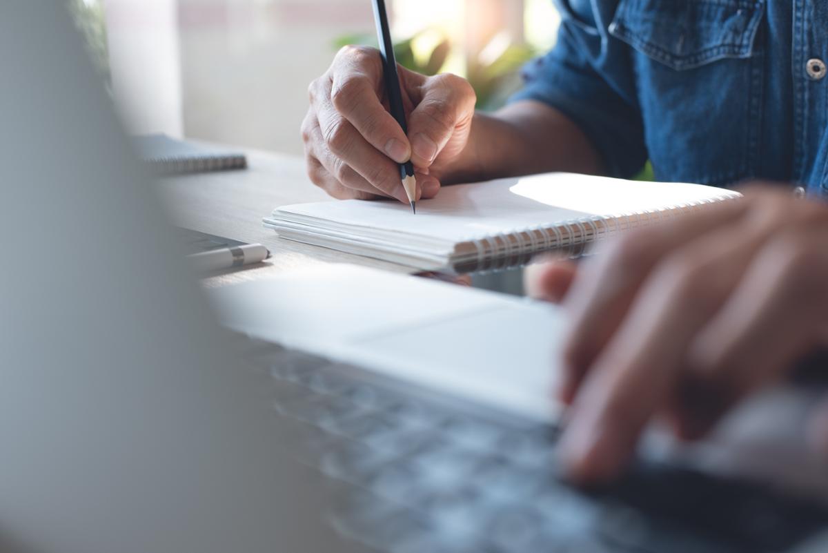 person writing on a notepad with one hand and typing on a laptop with another